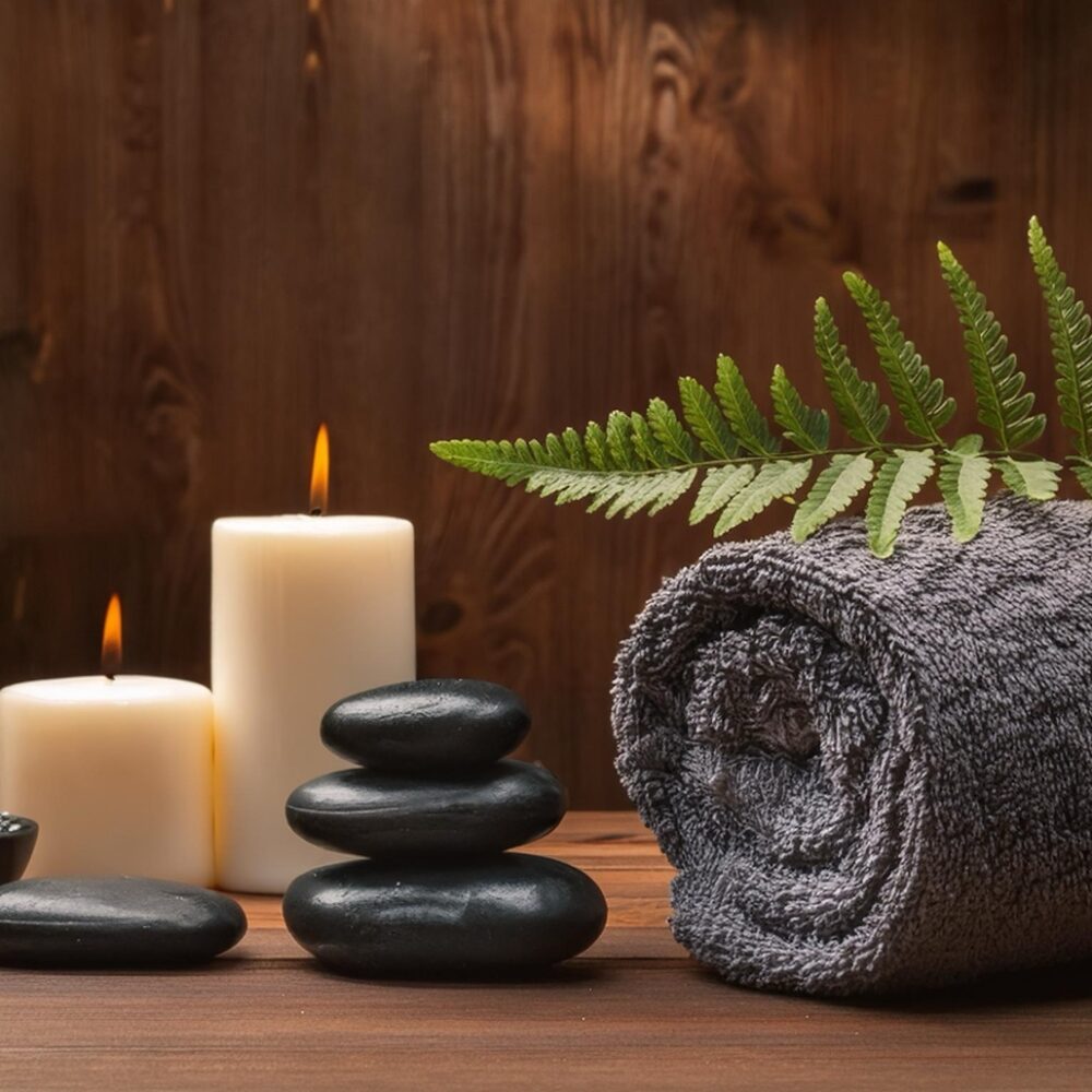 A spa setting featuring a gray towel rolled up beside lit candles and smooth stones. Fern leaves and a small bowl of black pebbles add to the serene atmosphere, set against a warm wooden background.